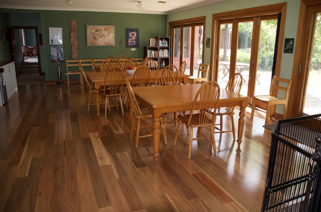 A dining room with wooden tables and chairs, large sliding glass door, and bookshelf against a green wall, with artwork and decorations.
