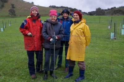 Four people dressed in colorful jackets and hats standing outdoors on a grassy hillside with a fence and trees in the background.