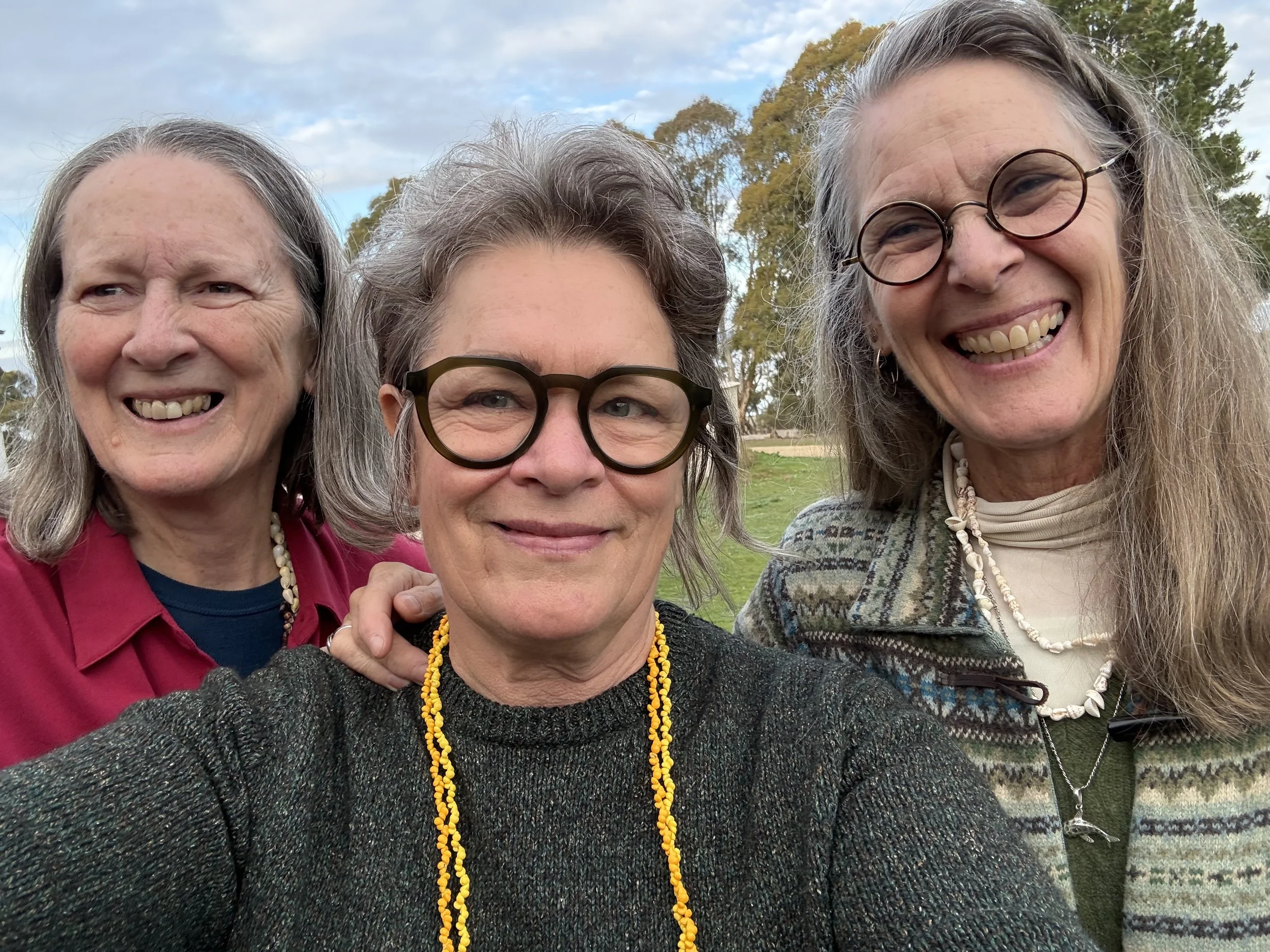 Three smiling women with glasses, long hair, and necklaces taking a selfie outdoors with trees and a partly cloudy sky in the background.