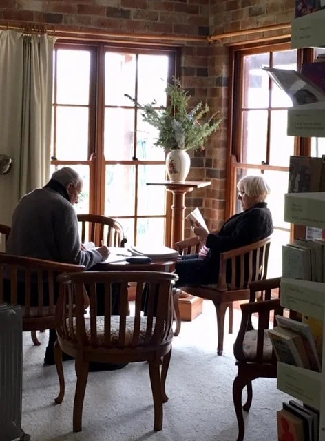 An elderly man and woman sitting in wooden chairs near large windows, reading and writing inside a brick-walled room with natural light, a tall vase with greenery on a pedestal between them.