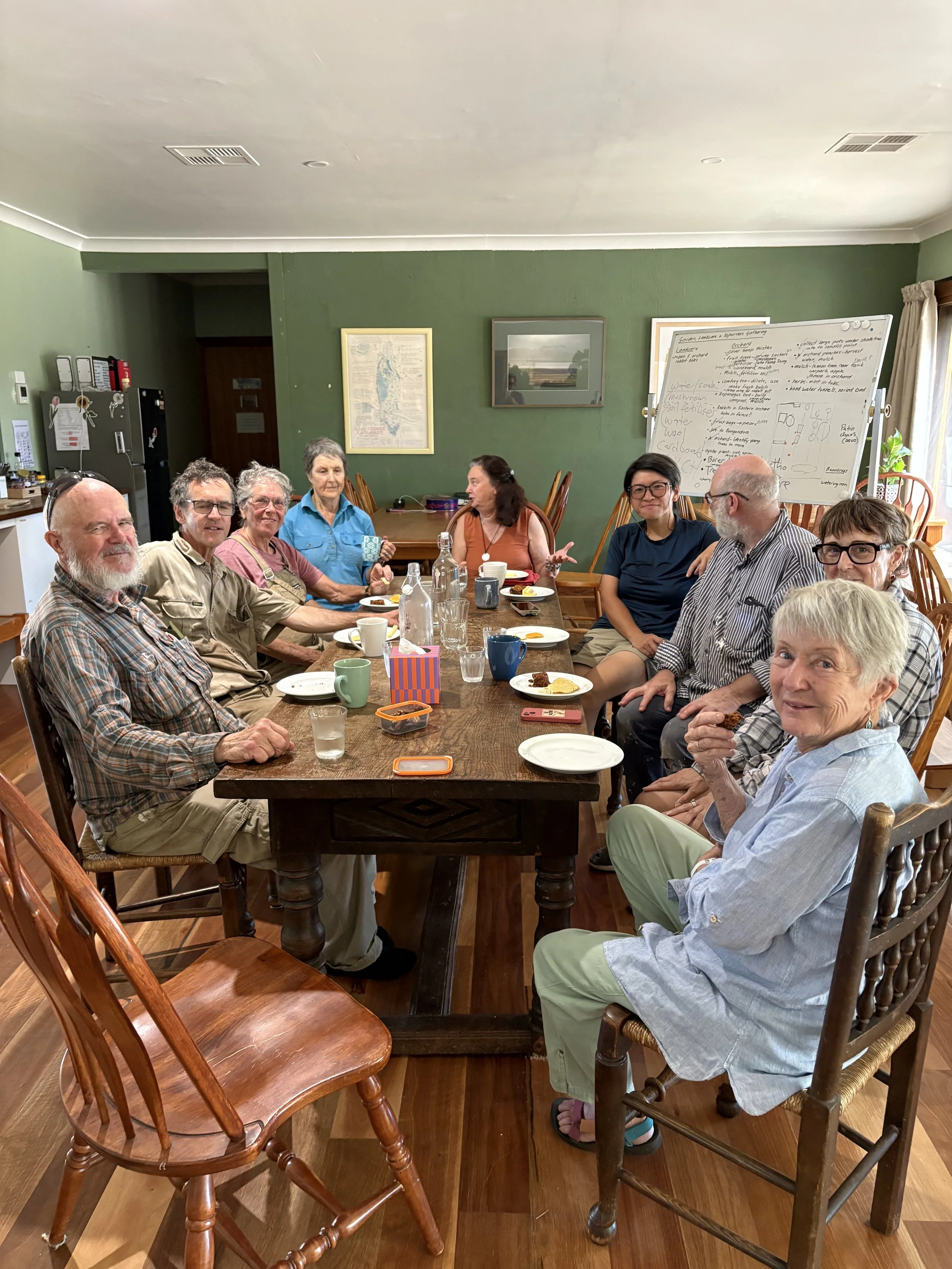 A group of nine people, mostly older adults, sitting around a dining table in a well-lit room, eating dessert and chatting. The room has green walls, a whiteboard with notes, and a framed picture. There are various mugs, plates, and water bottles on the table.