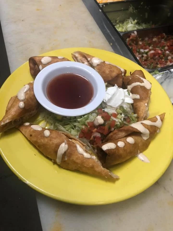 Plate of golden brown taquitos topped with white sauce, served with shredded lettuce, diced tomatoes, sour cream, and a side of red sauce in a small bowl.