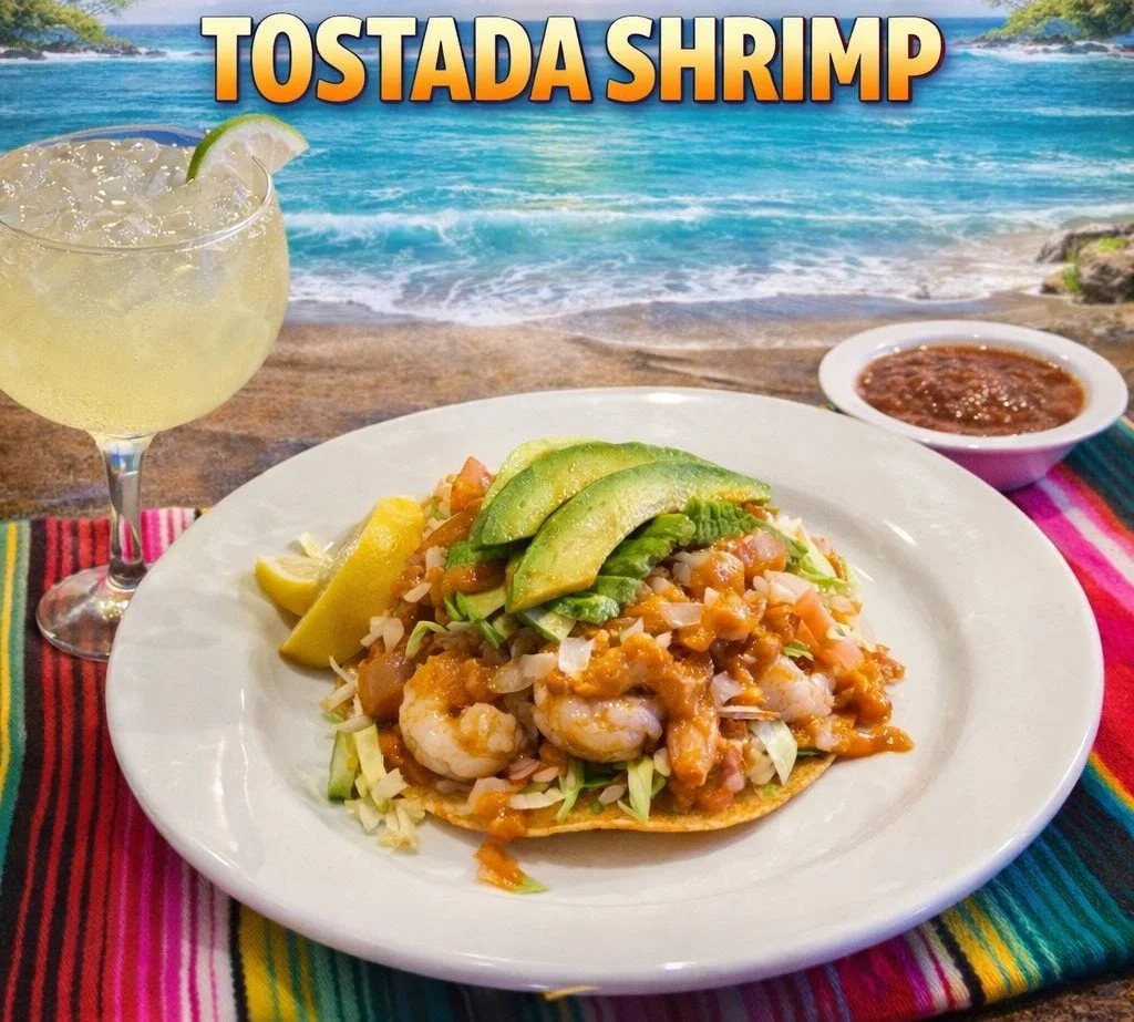 A plate of shrimp tostada with avocado slices and shredded lettuce, accompanied by a lemon wedge, beside a small bowl of salsa, on a colorful striped tablecloth with a beach and ocean in the background and a cocktail with lime in a glass.
