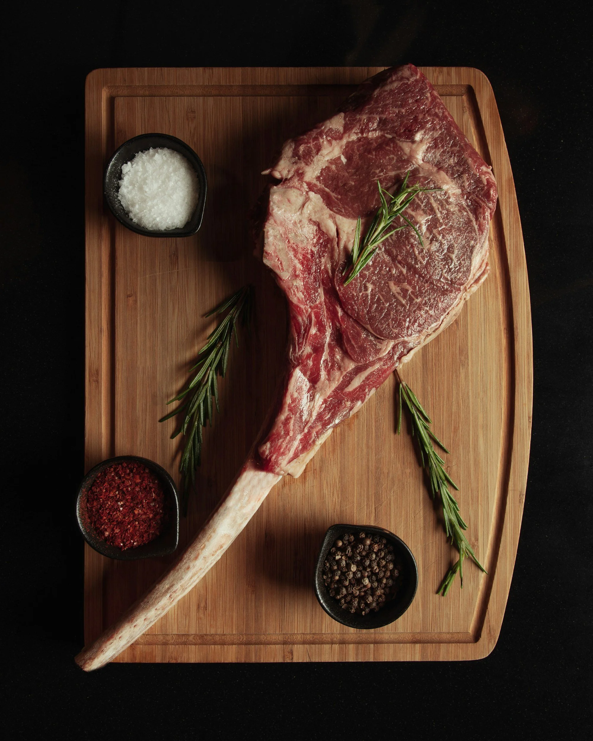 Raw ribeye steak on a wooden cutting board, garnished with rosemary sprigs and surrounded by bowls of sea salt, red pepper flakes, and black peppercorns.