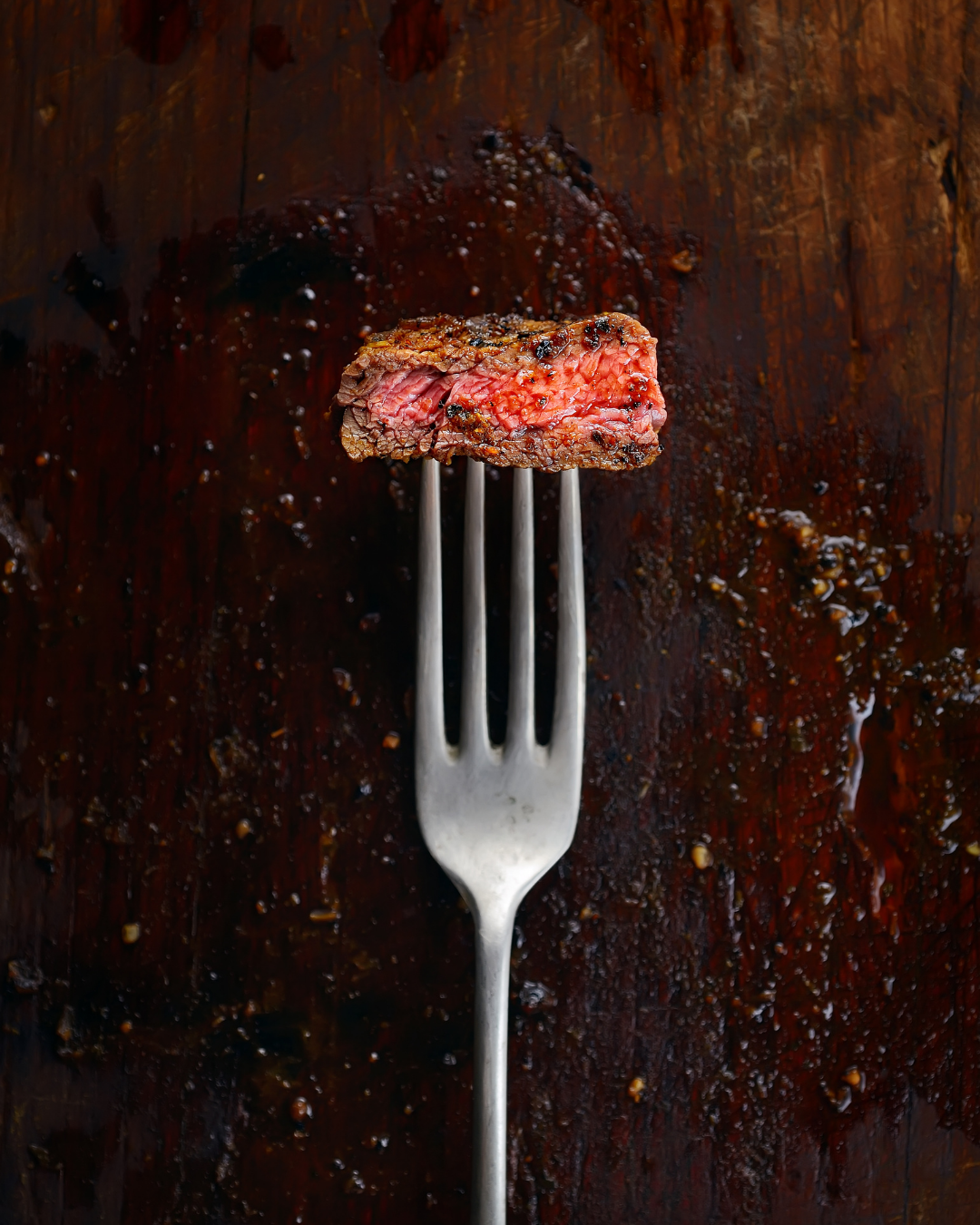 A piece of cooked steak on a fork over a dark wooden surface.