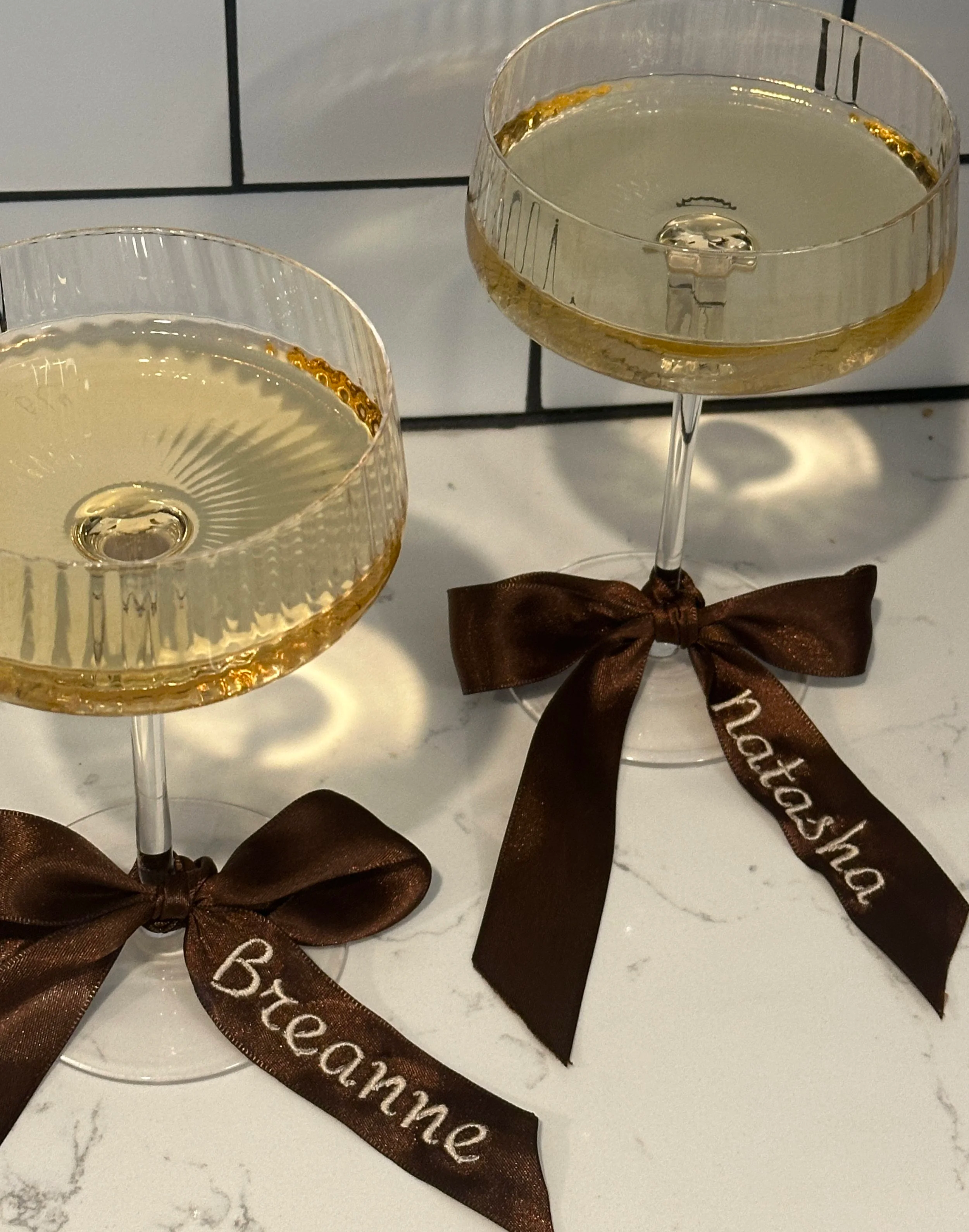 Two elegant champagne glasses filled with champagne, decorated with brown ribbons that have white embroidered text reading ‘Breanne’ and ‘Natasha’ as bridesmaids gifts.