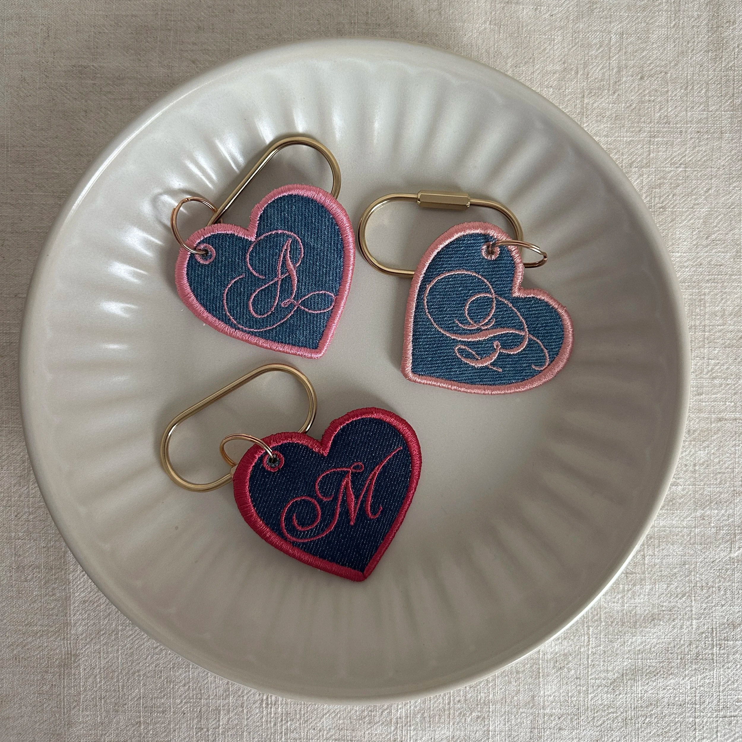 Three embroidered denim heart-shaped keychains with cursive initials, on a white ceramic plate.