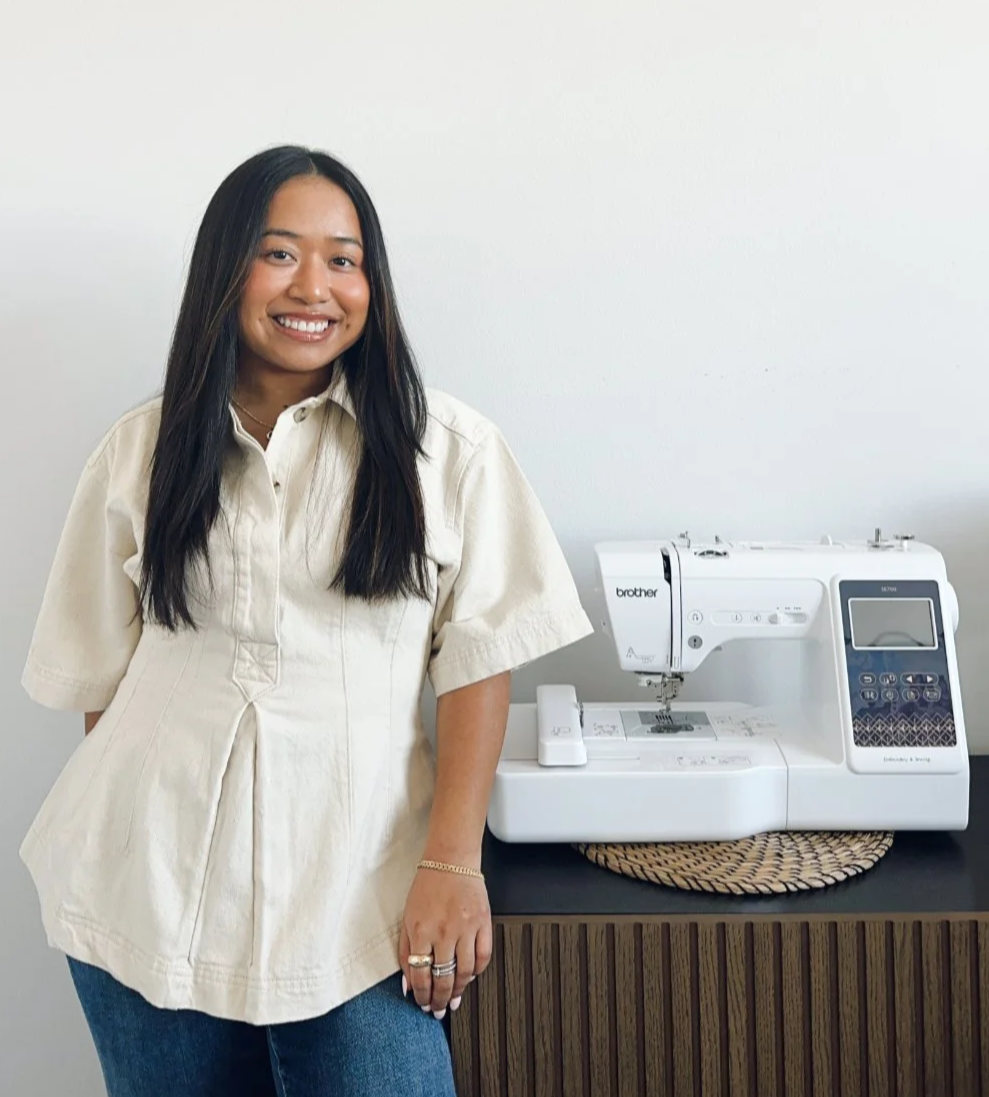 Founder of Dream Girl standing next to a white embroidery sewing machine on a black table with a woven mat underneath, against a plain white wall.