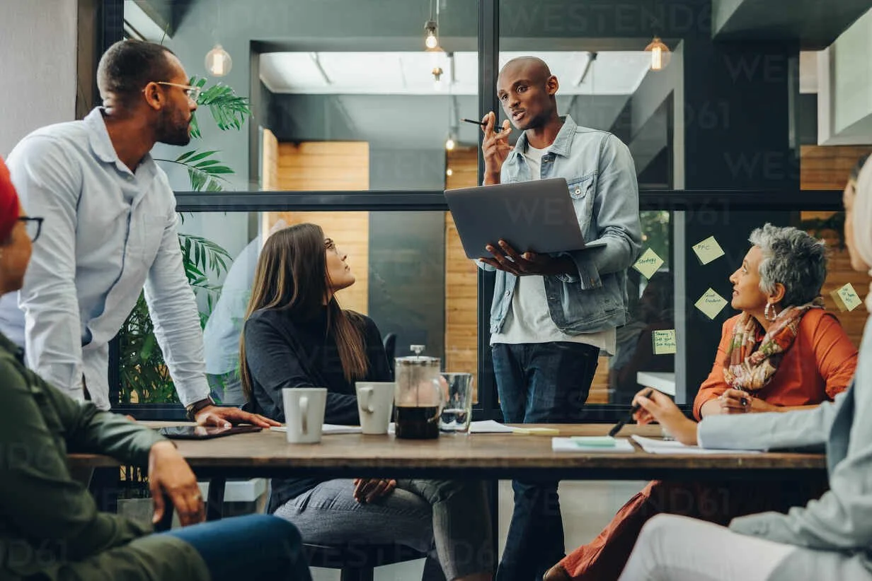 A diverse group of people having a discussion in a modern office conference room, with one person standing and speaking while others are seated around a table.