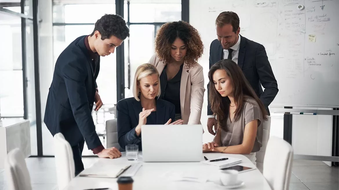 Group of five diverse business people gathered around a laptop in a modern conference room, collaborating on a project.