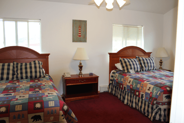 Two beds with checkered and patterned bedspreads, nightstand with lamp and phone, and a picture of a lighthouse on the wall in a bedroom.