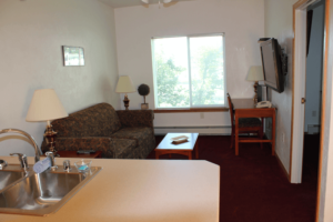 Living room with a floral-patterned sofa, two table lamps, a wooden side table, a window with a view of trees, a wall-mounted TV, and a doorway.