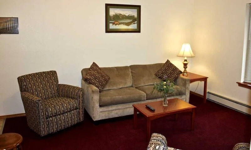 Living room with a beige sofa, patterned armchair, wooden coffee table with a remote and flower vase, side table with lamp, framed landscape picture, and window with blinds.