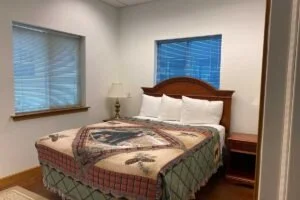 A bedroom with two windows, a bed with a patterned quilt, white pillows, a wooden headboard, a nightstand, and a lamp.