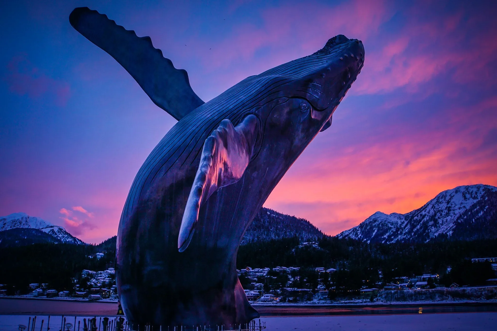 Large whale sculpture illuminated at dusk, with a mountain range and a small town in the background.