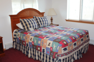 A neatly made bed with a colorful patchwork quilt and plaid pillows in a bedroom with white walls, a wooden headboard, a nightstand with a lamp, and a window with white curtains.