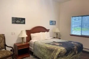 A bedroom with a wooden bed frame, white bedding, two matching lamps on wooden nightstands, a window with blinds, and a chair with patterned upholstery.