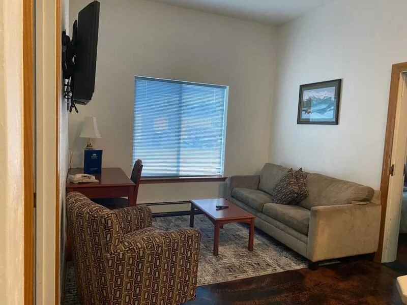 Living room with a beige sofa, patterned armchair, wooden coffee table, window with closed blinds, wall-mounted TV, side table with lamp and phone, framed picture on wall, and doorway.