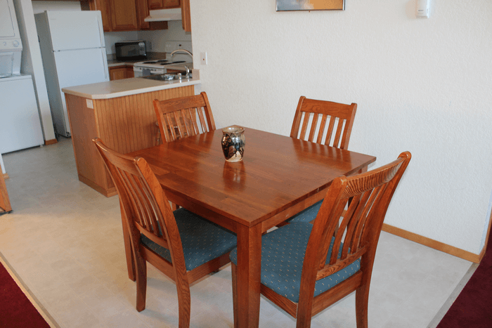 A wooden dining table with four chairs, a ceramic vase, in a room with a kitchen in the background.