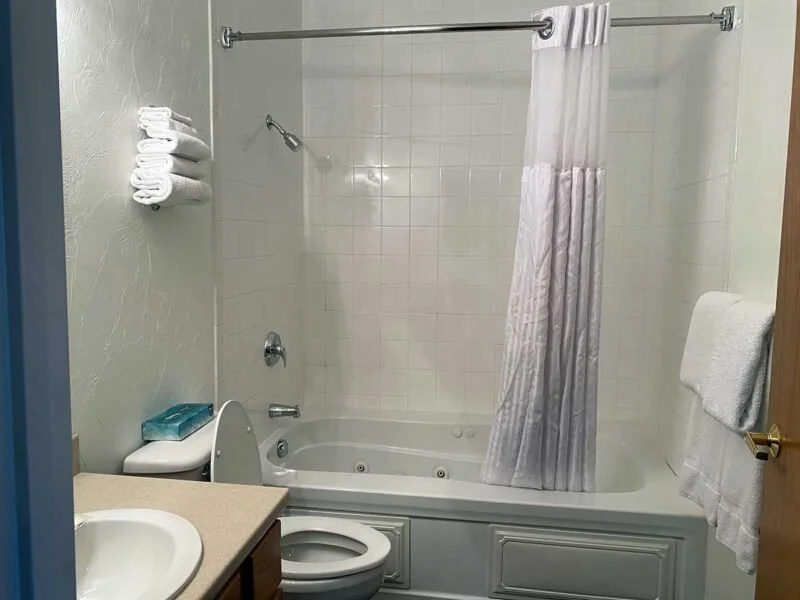 Bathroom with bathtub, shower, and white tiled walls, a towel rack with folded towels, a toilet, a small sink with a countertop, and a shower curtain.