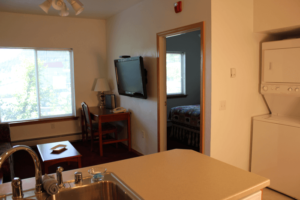 Interior view of a small apartment or hotel room with a sink and kitchen area, a window, a dining table with a chair, a wall-mounted TV, and a doorway leading to a bedroom.