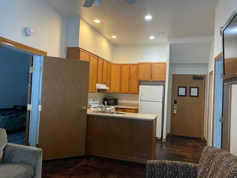 Interior view of a kitchen in a hotel room or apartment with wooden cabinets, white refrigerator, microwave, and a small breakfast bar. Part of a living area with armchairs and a doorway leading to another room is visible.