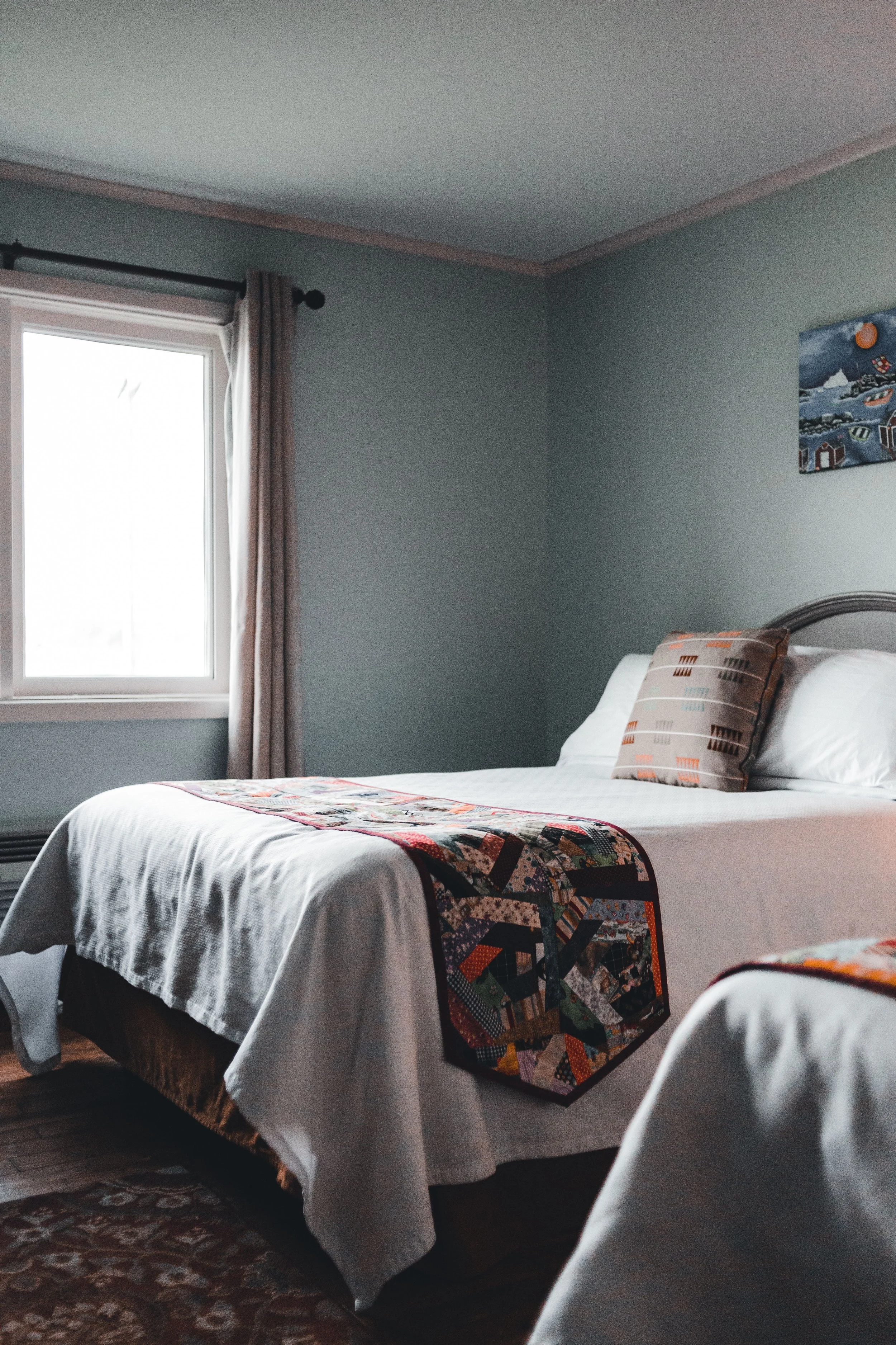 A bedroom with two beds, each covered with white sheets and a colorful patchwork quilt at the foot. One bed has a decorative pillow. There is a window with curtains on the left and framed artwork on the light green wall to the right.