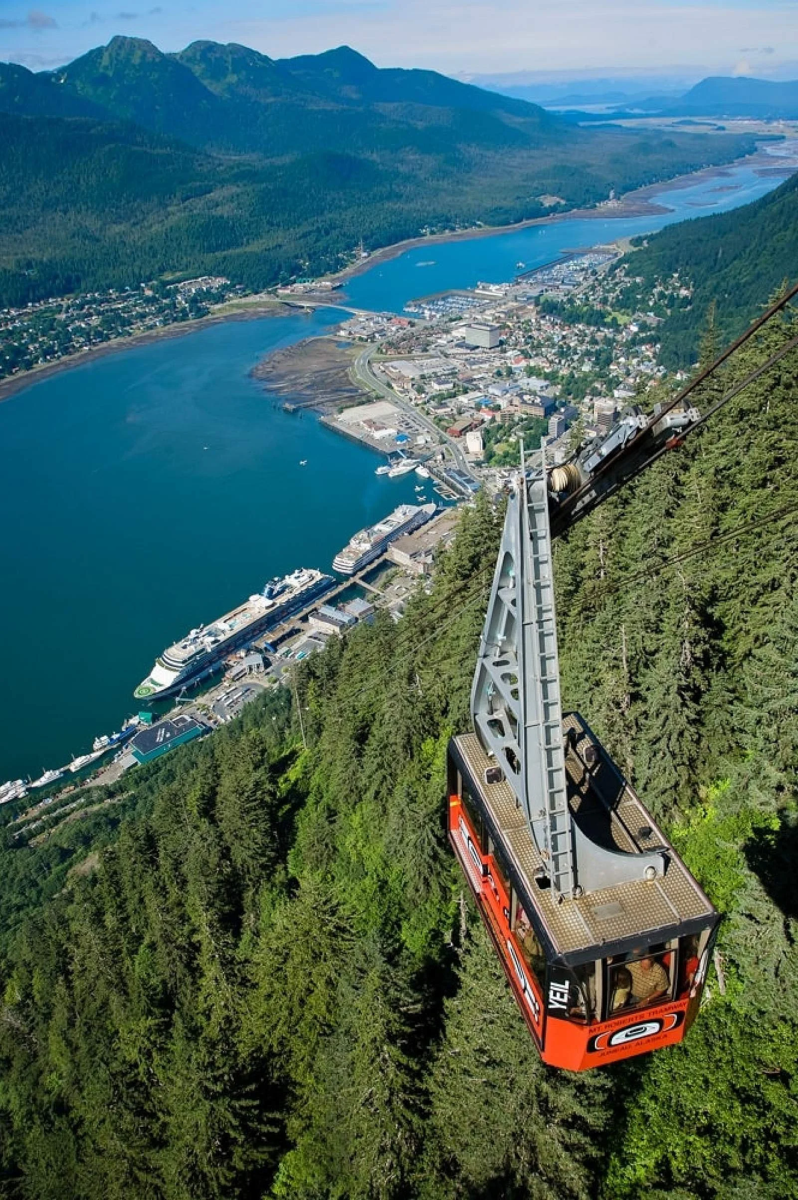 A scenic aerial view of a city by a large river, surrounded by lush green forests and mountains, with a cable car ascending a mountainside in the foreground.