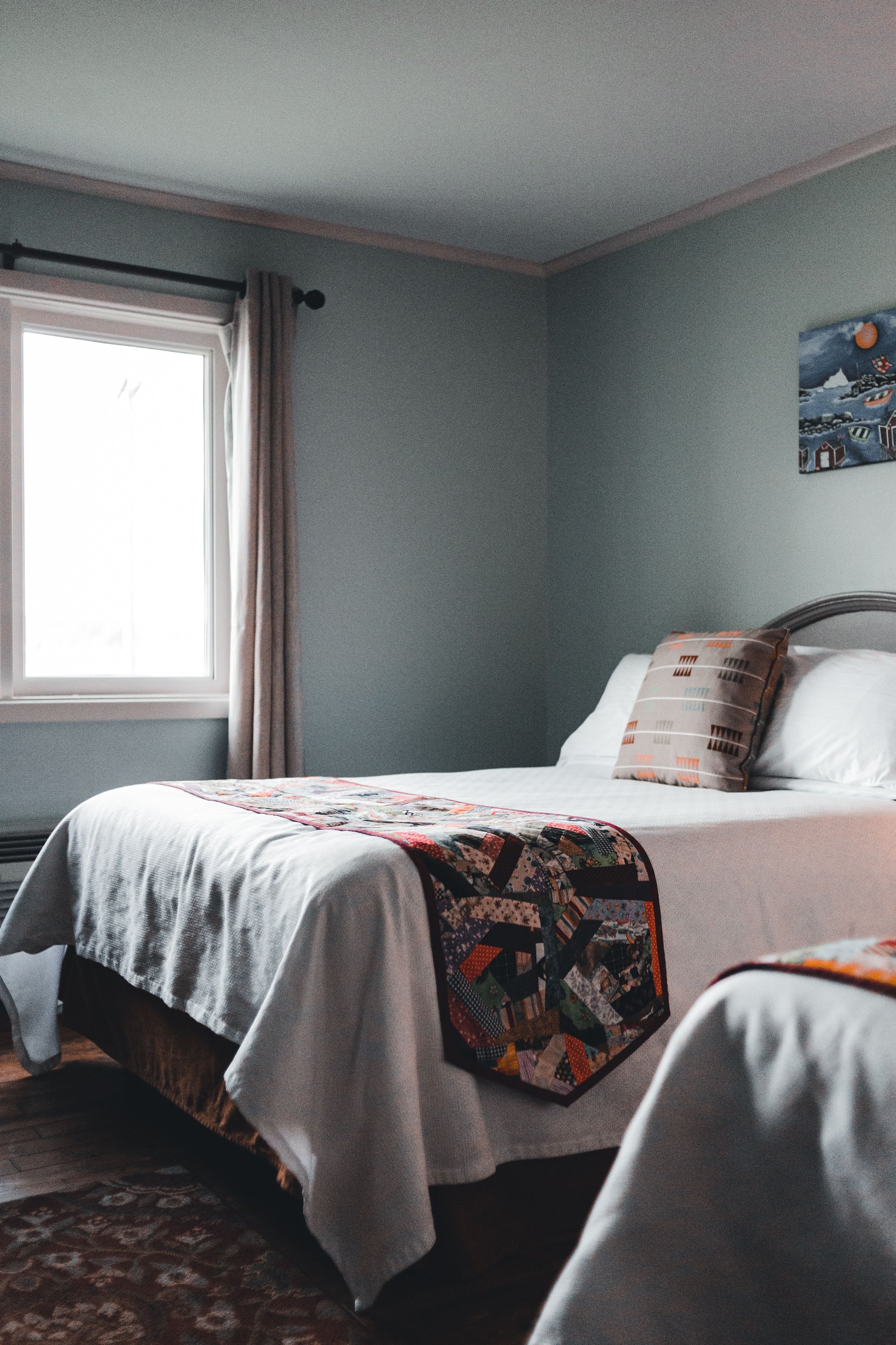 A cozy bedroom with a bed covered in white linens, a colorful quilt at the foot, a plaid pillow, a window with beige curtains, and a landscape-themed painting on the wall.