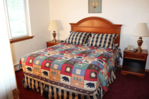 A cozy bedroom with a wooden bed frame and a colorful patchwork quilt featuring bear patterns. There are two nightstands with lamps on either side of the bed, and a window with green foliage outside.