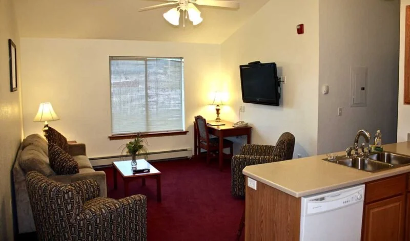 Living room with a couch, two armchairs, a coffee table, a TV on the wall, and a window with blinds, connecting to a kitchen with a sink and dishwasher.