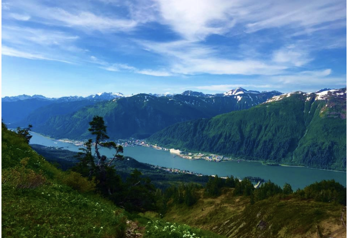 Scenic view of a river flowing through lush green mountains with snow-capped peaks under a partly cloudy blue sky.