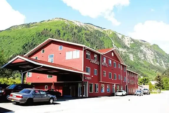 Red hotel building with a parking lot and cars, set against green mountains under a partly cloudy sky.