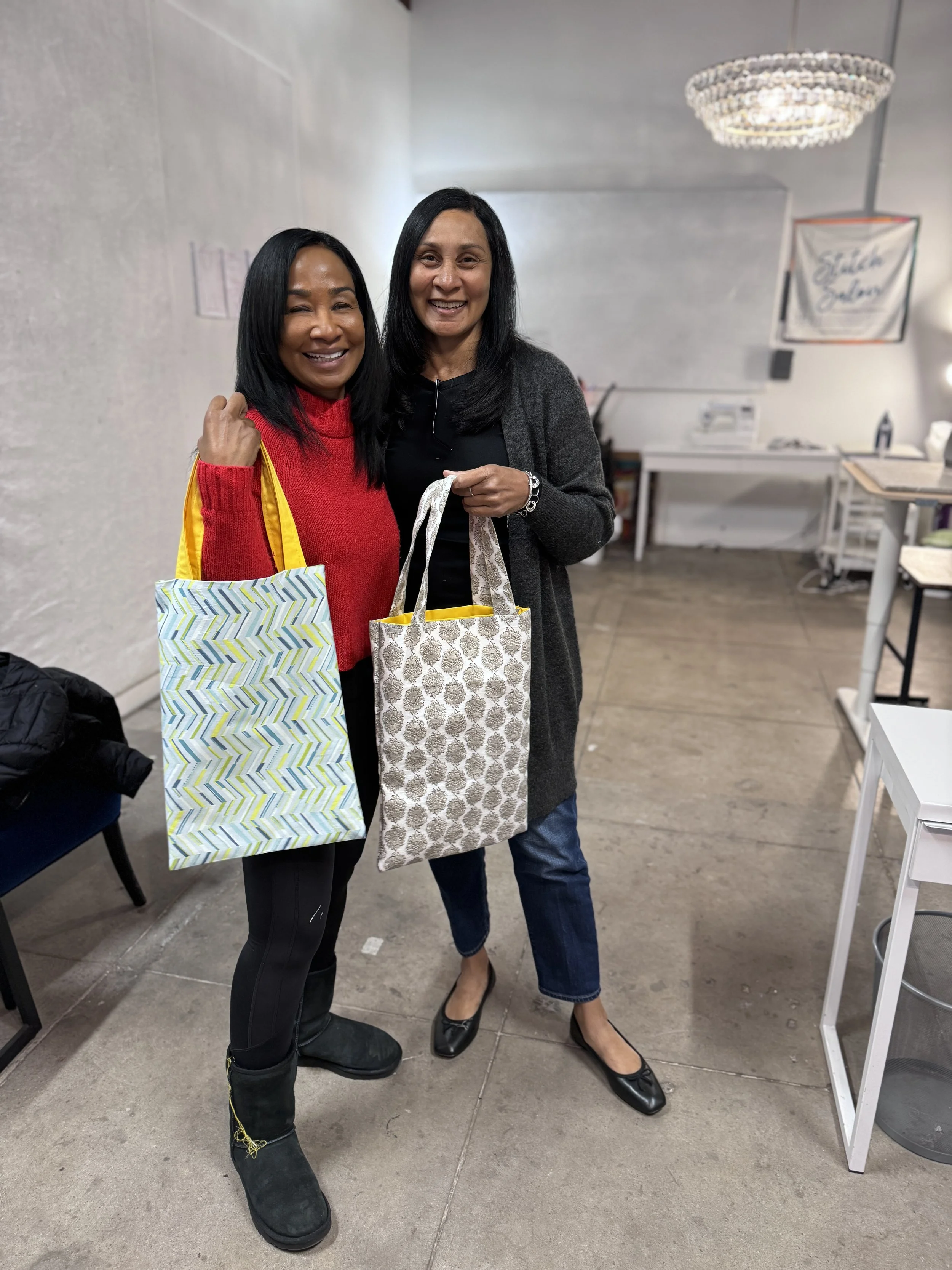 Two women standing indoors, smiling, holding handmade tote bags, with a chandelier and a sign in the background.