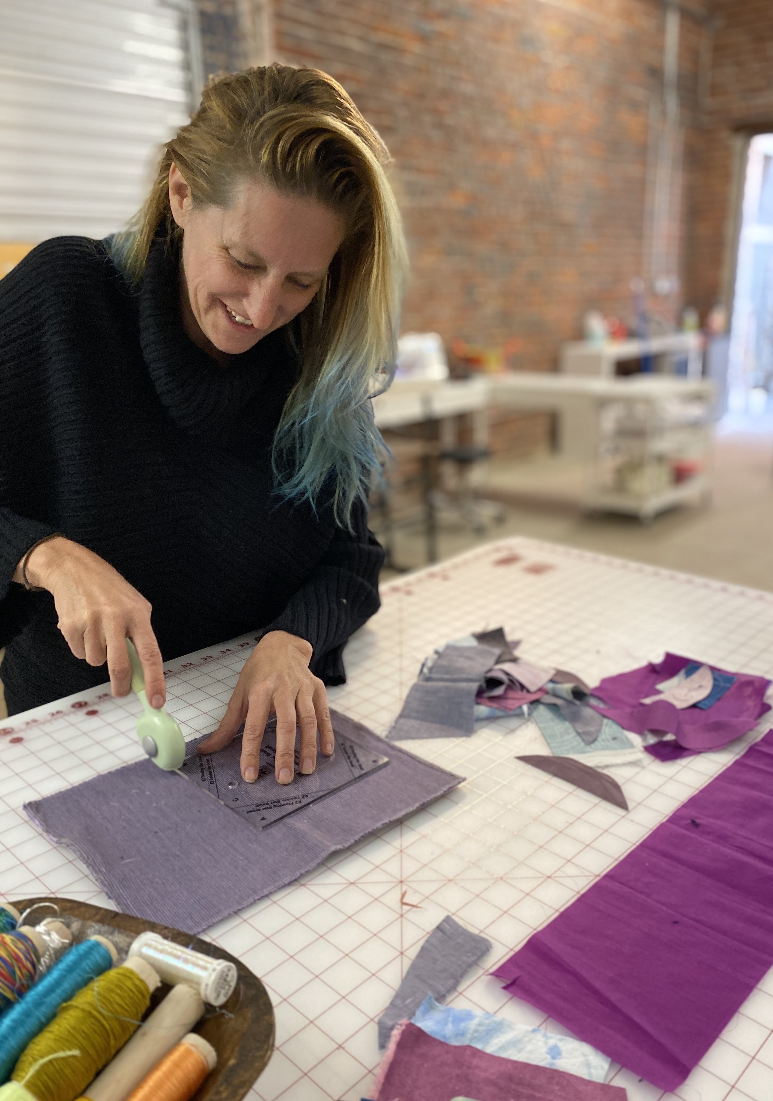 Woman using a rotary cutter to cut fabric on a worktable with various fabric scraps and sewing supplies in a craft room.