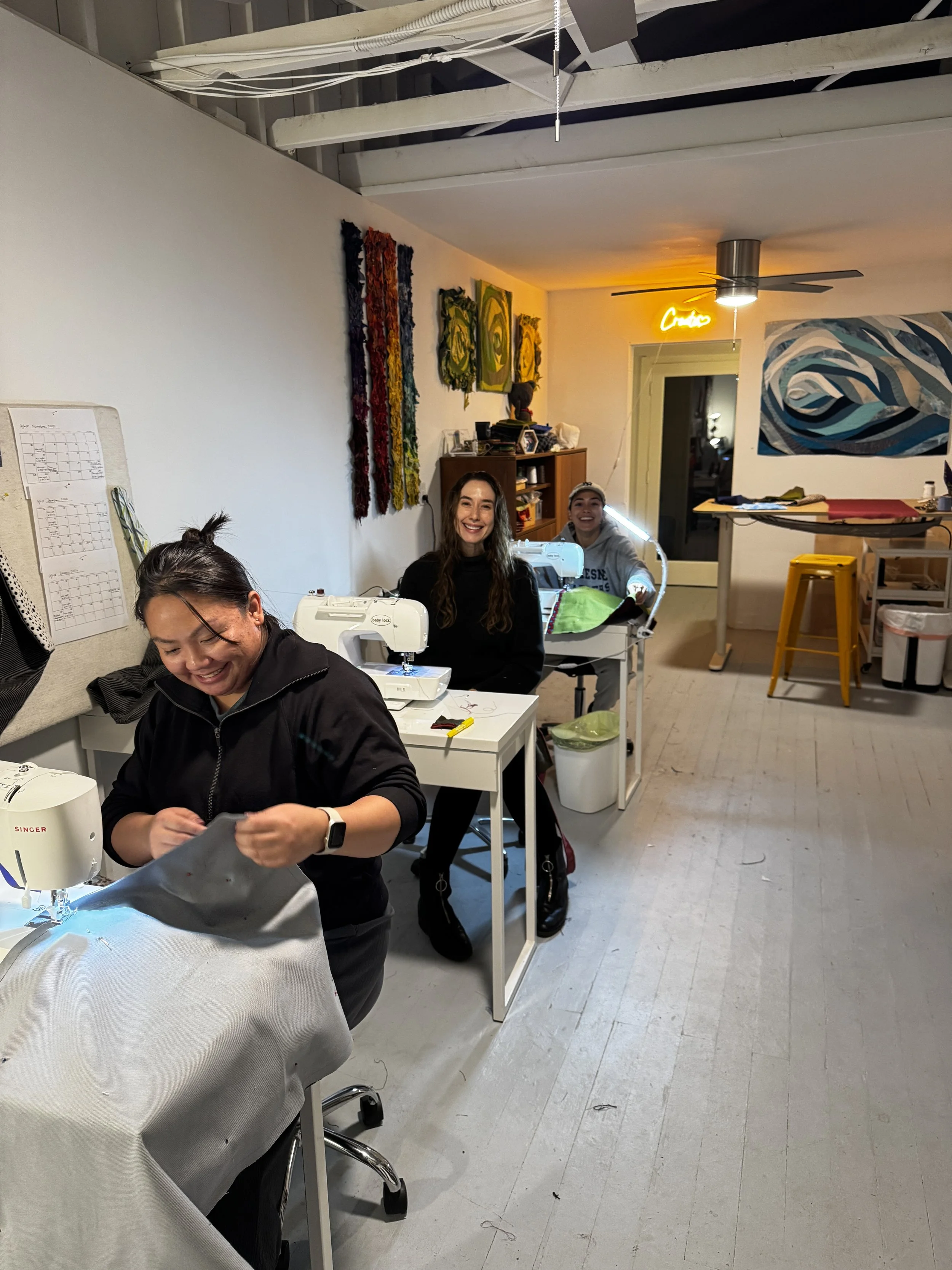 Three women working in a craft room with sewing machines, colorful wall art, and a neon sign that says 'Cactus' in the background.