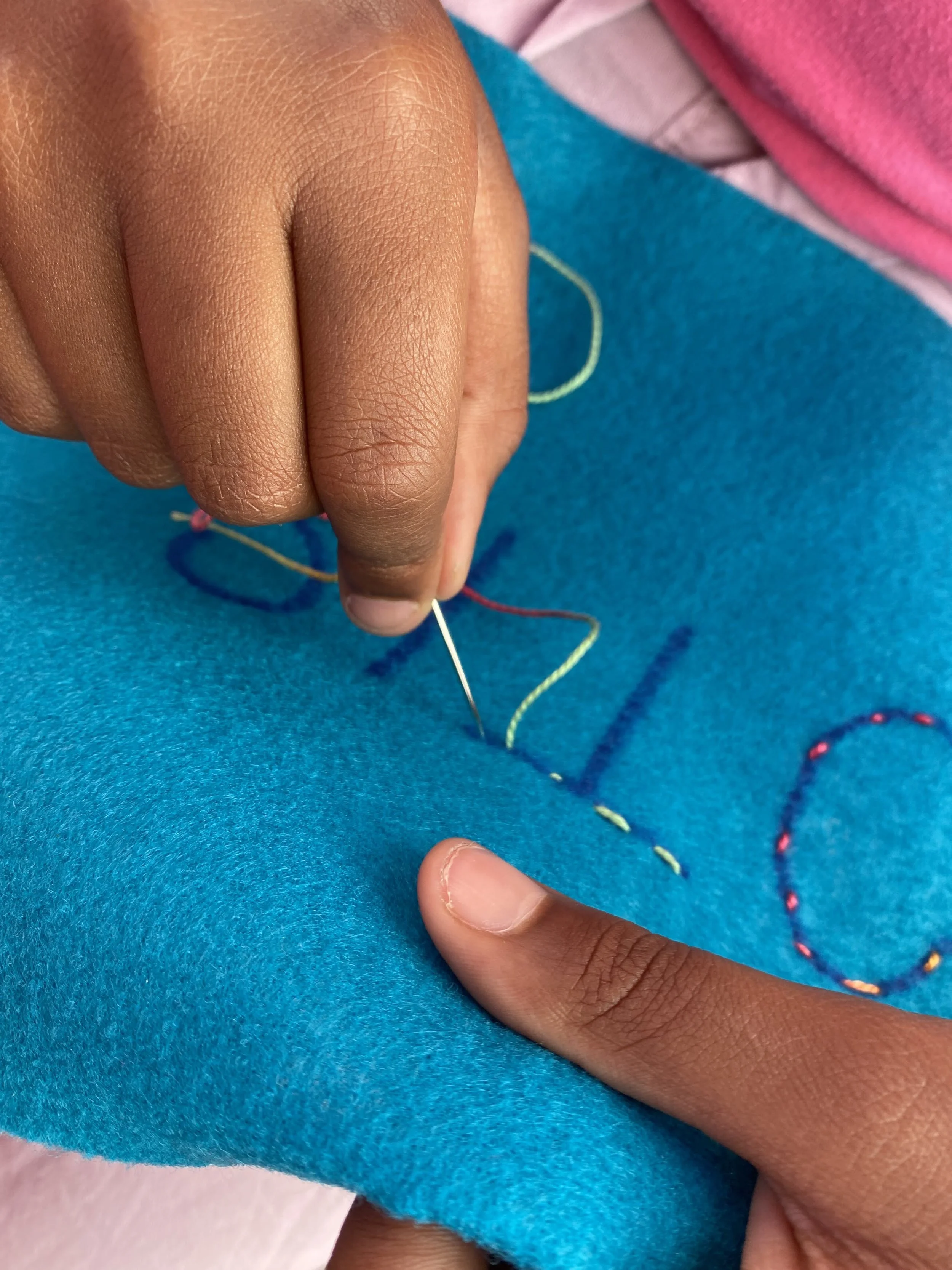 Close-up of a person sewing with a needle and colorful thread on a blue fabric.