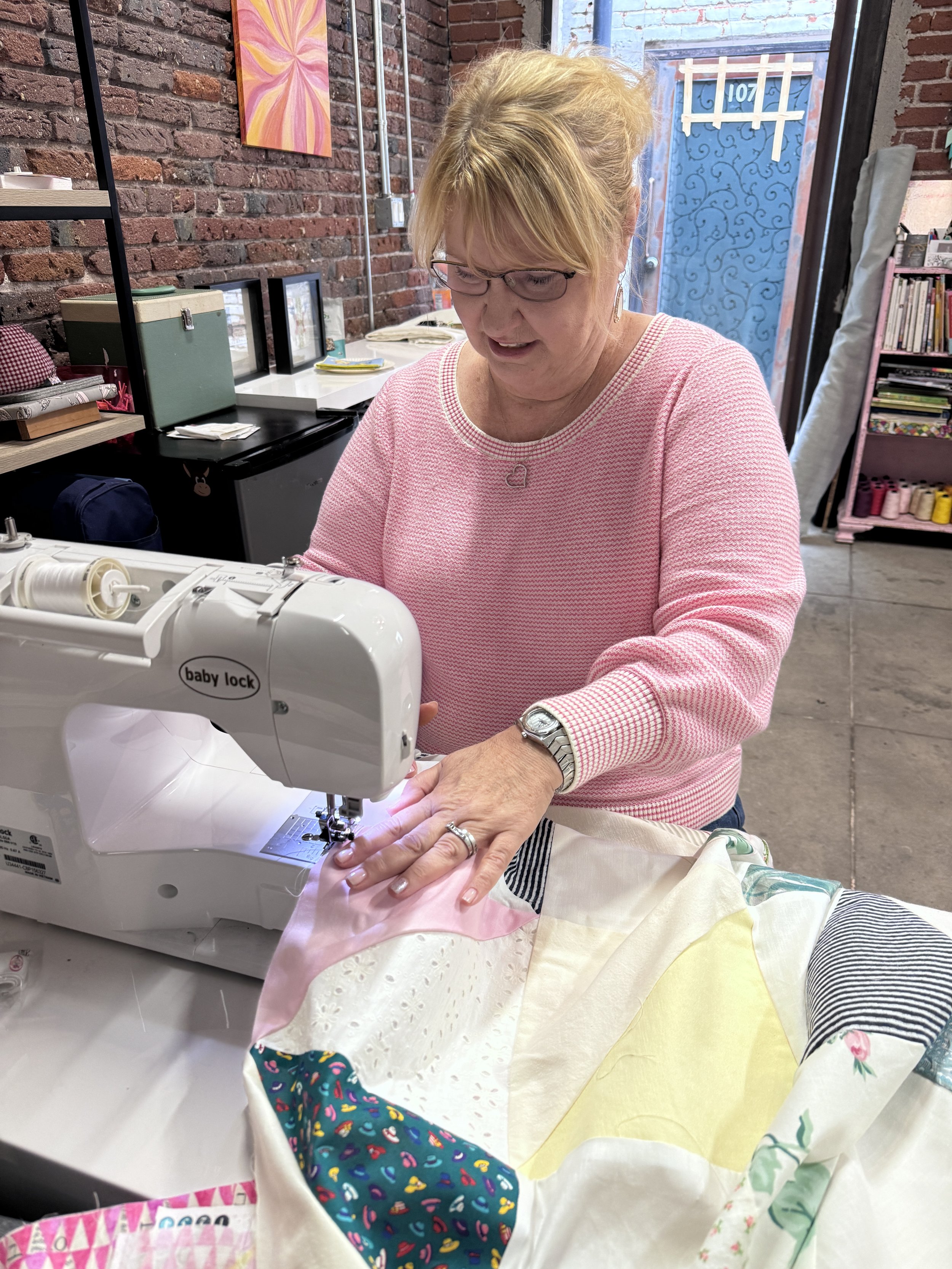 A woman sewing on a Baby Lock sewing machine in a sewing classroom with artwork, and shelves of supplies.