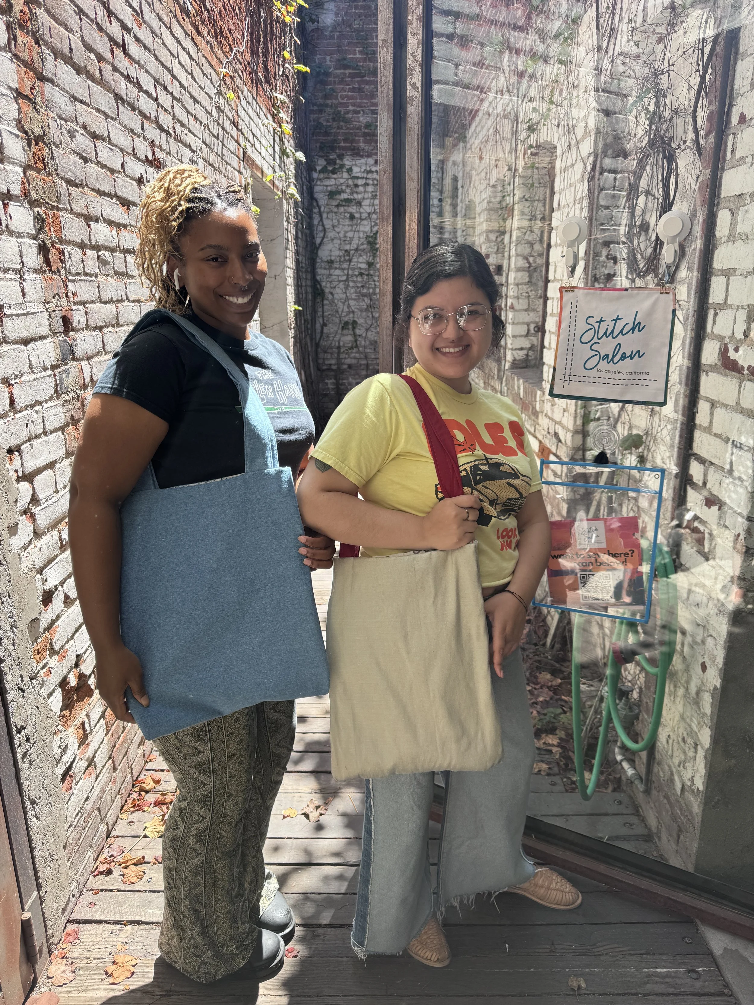 Two women standing outside a shop called Stitch Salon, with brick walls and a sign indicating Los Angeles, California. They are smiling, holding tote bags, with sunlight streaming through. The woman on the left has blond, curly hair and is wearing a 