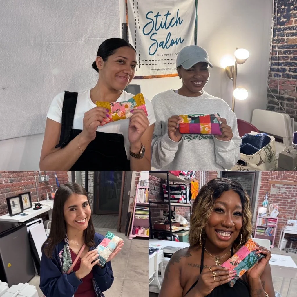 Four women indoors holding colorful fabric pouches or wallets, smiling at the camera. The background shows a sign for Stitch Salon, a brick wall, and shelves with books and decor.