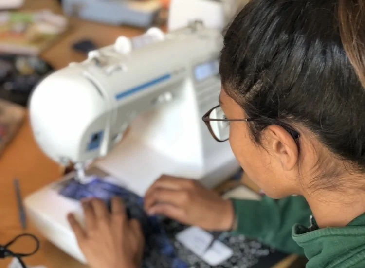 A woman with glasses sewing fabric with a sewing machine on a wooden table.