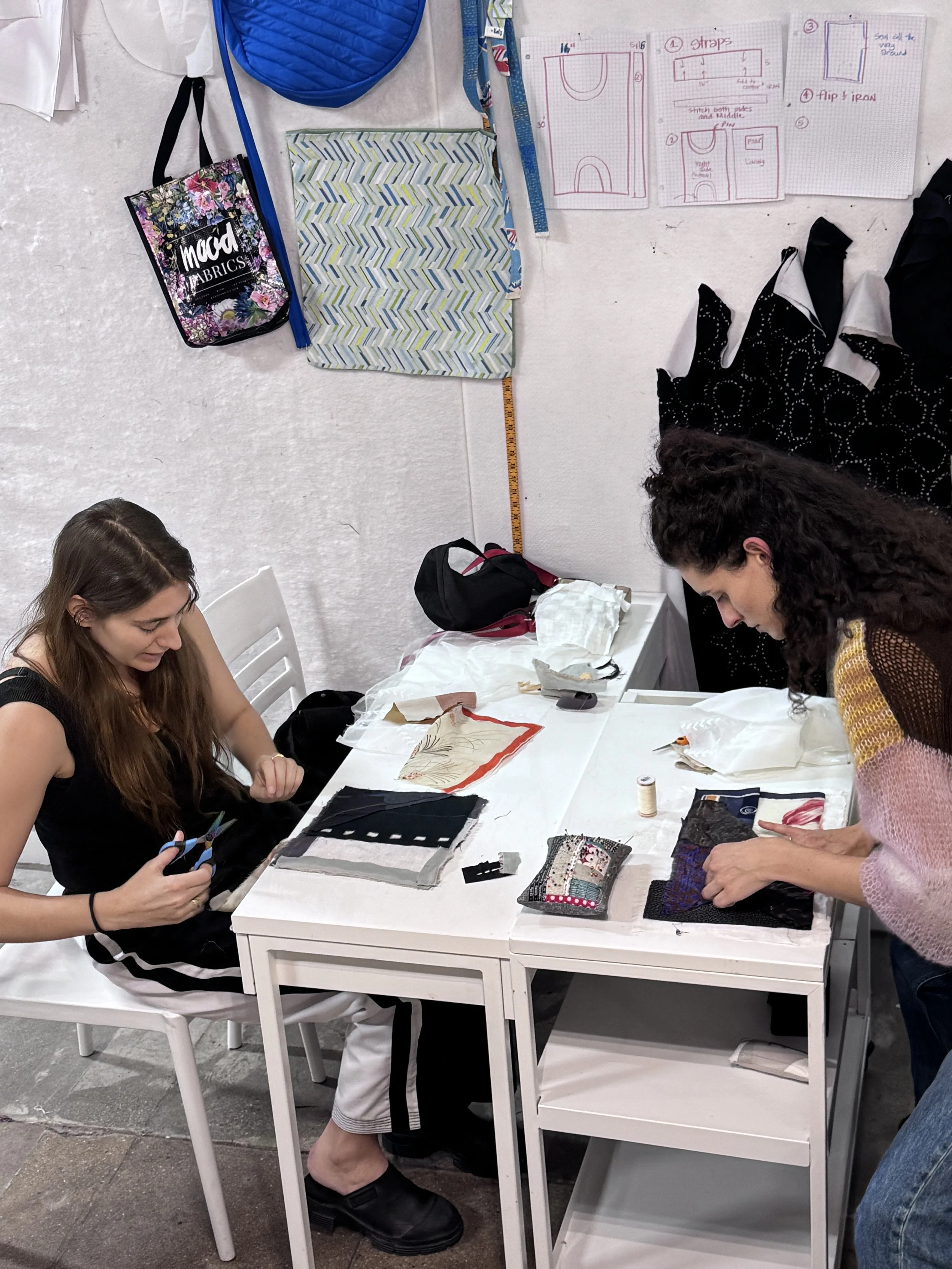 Two women working on sewing projects at a white table covered with fabric swatches, sewing supplies, and small projects, with sewing patterns and notes pinned to the wall behind them.