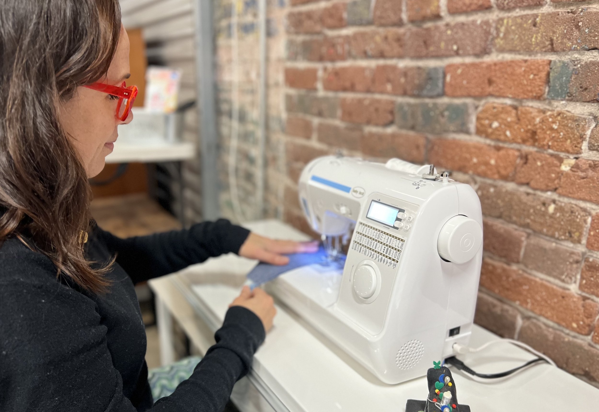 A woman with red glasses sewing with a sewing machine on a white table against an exposed brick wall.