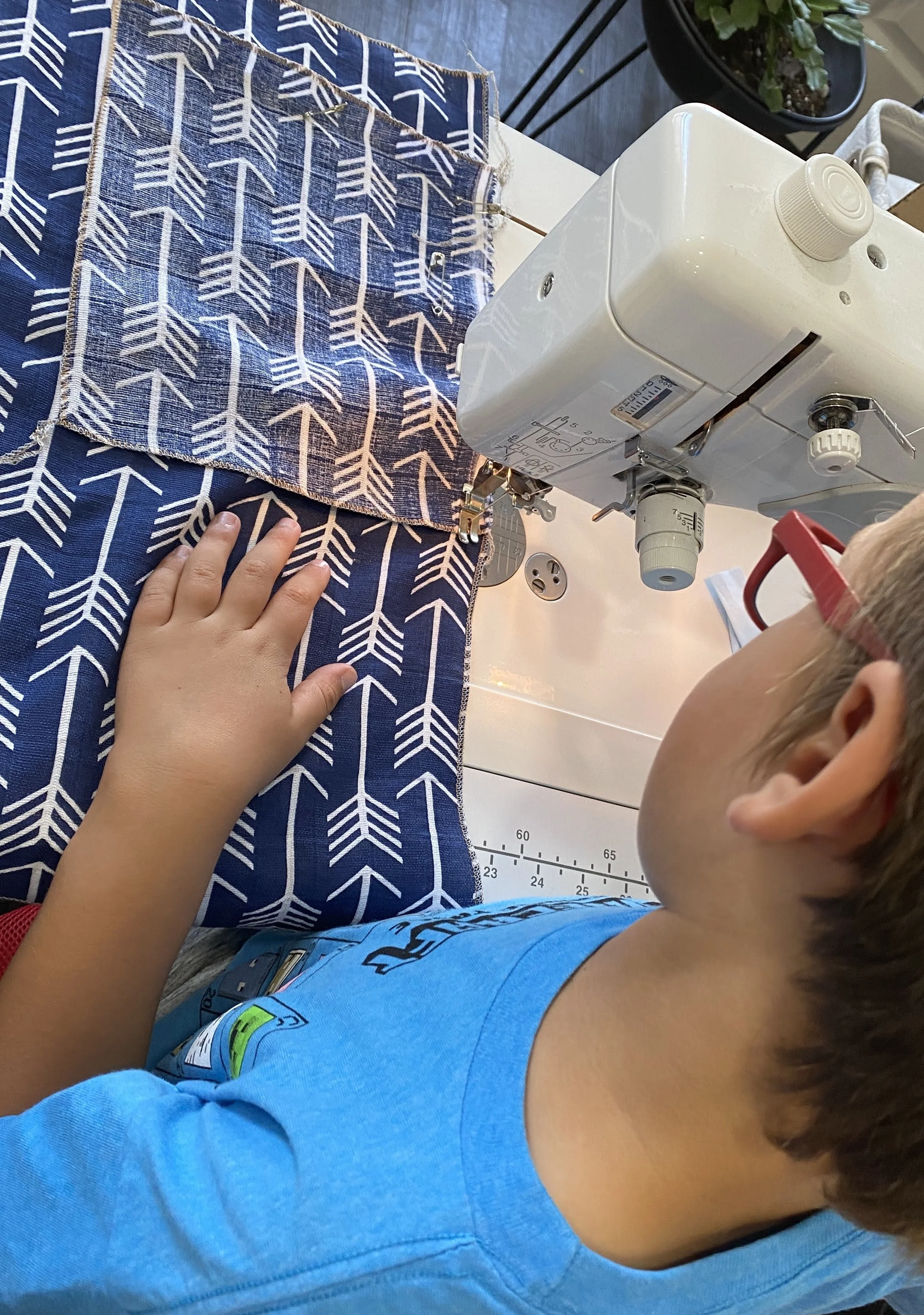 A young boy sewing fabric with a white sewing machine, wearing red glasses and a blue shirt, working on two pieces of fabric with a navy blue and white arrow pattern.