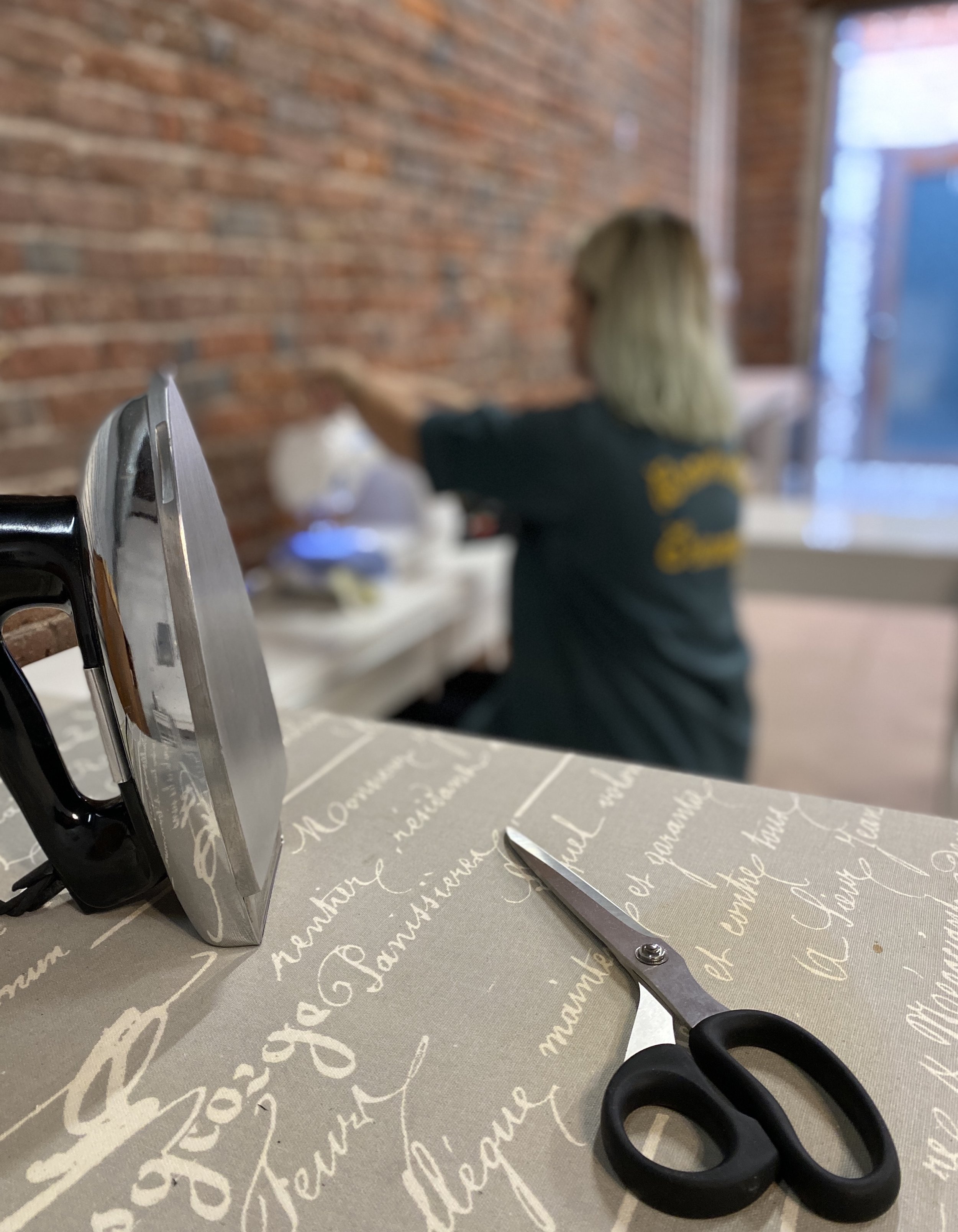 A pair of scissors and an iron on a table with a tablecloth, with a woman in the background working at a table against a brick wall.