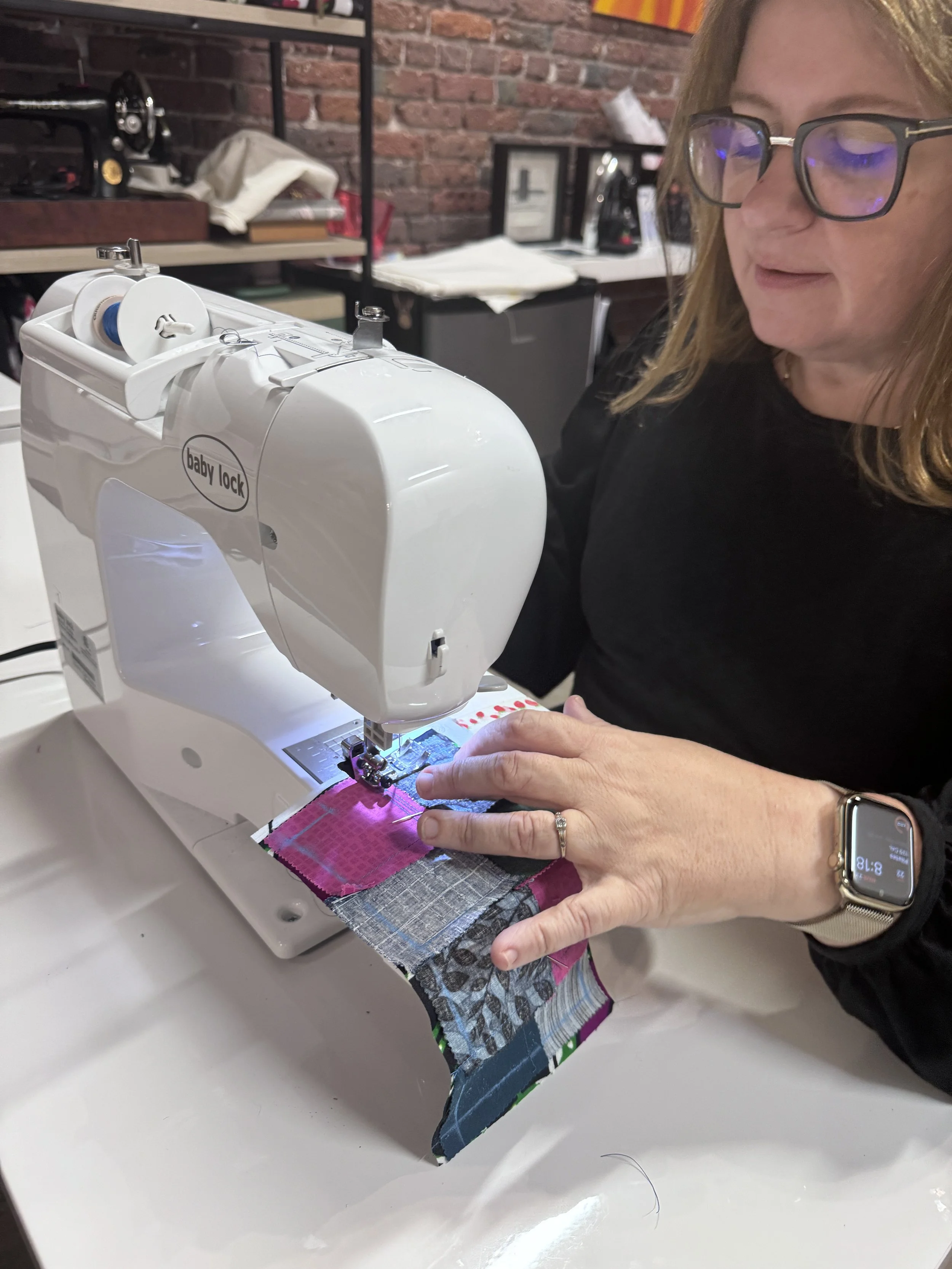 Woman sewing colorful fabric on a Baby Lock sewing machine in a room with brick walls and sewing supplies.