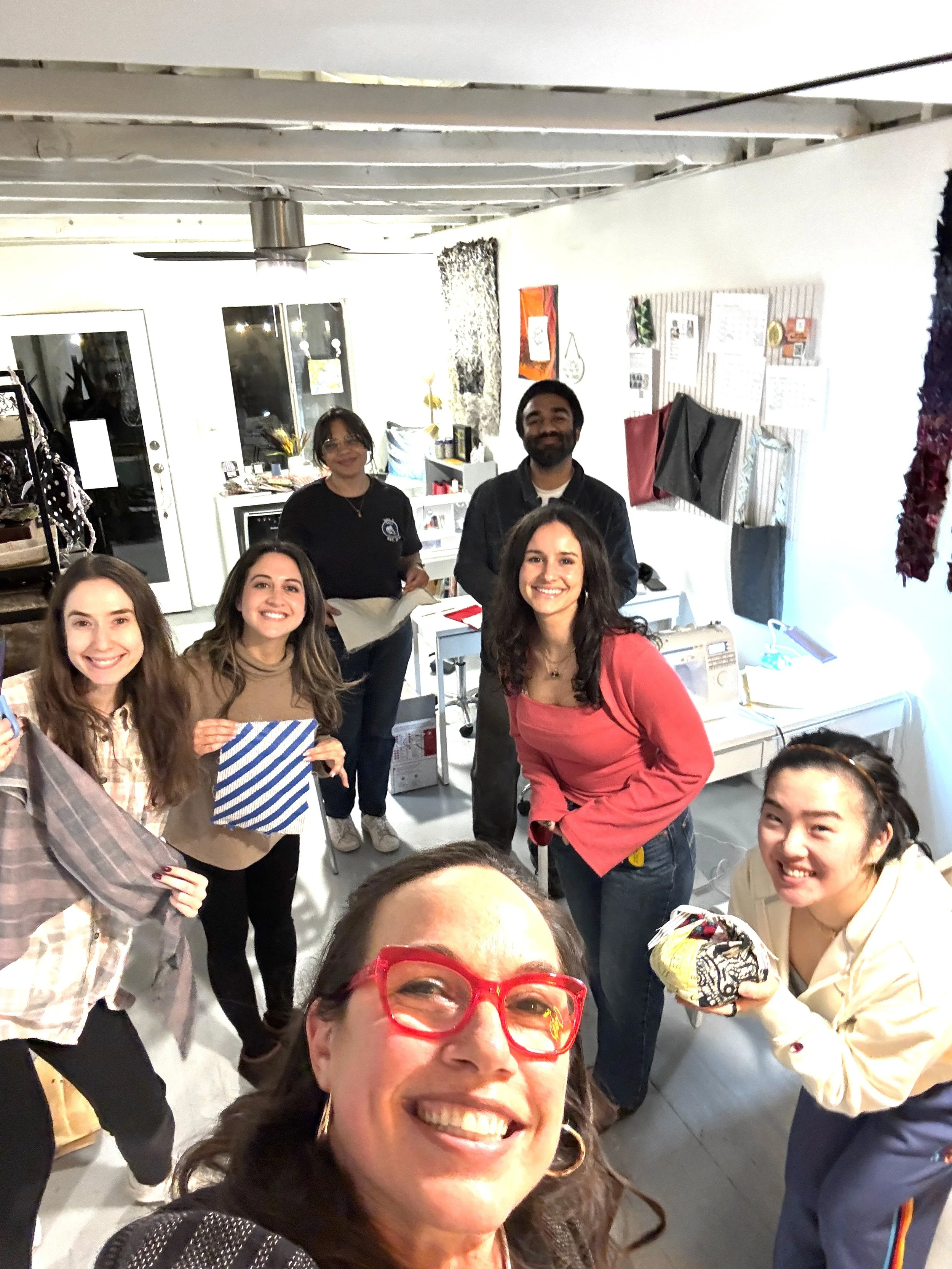 Group of seven people smiling and posing for a photo in an indoor space, with at least one person holding a piece of fabric, and others holding various items.