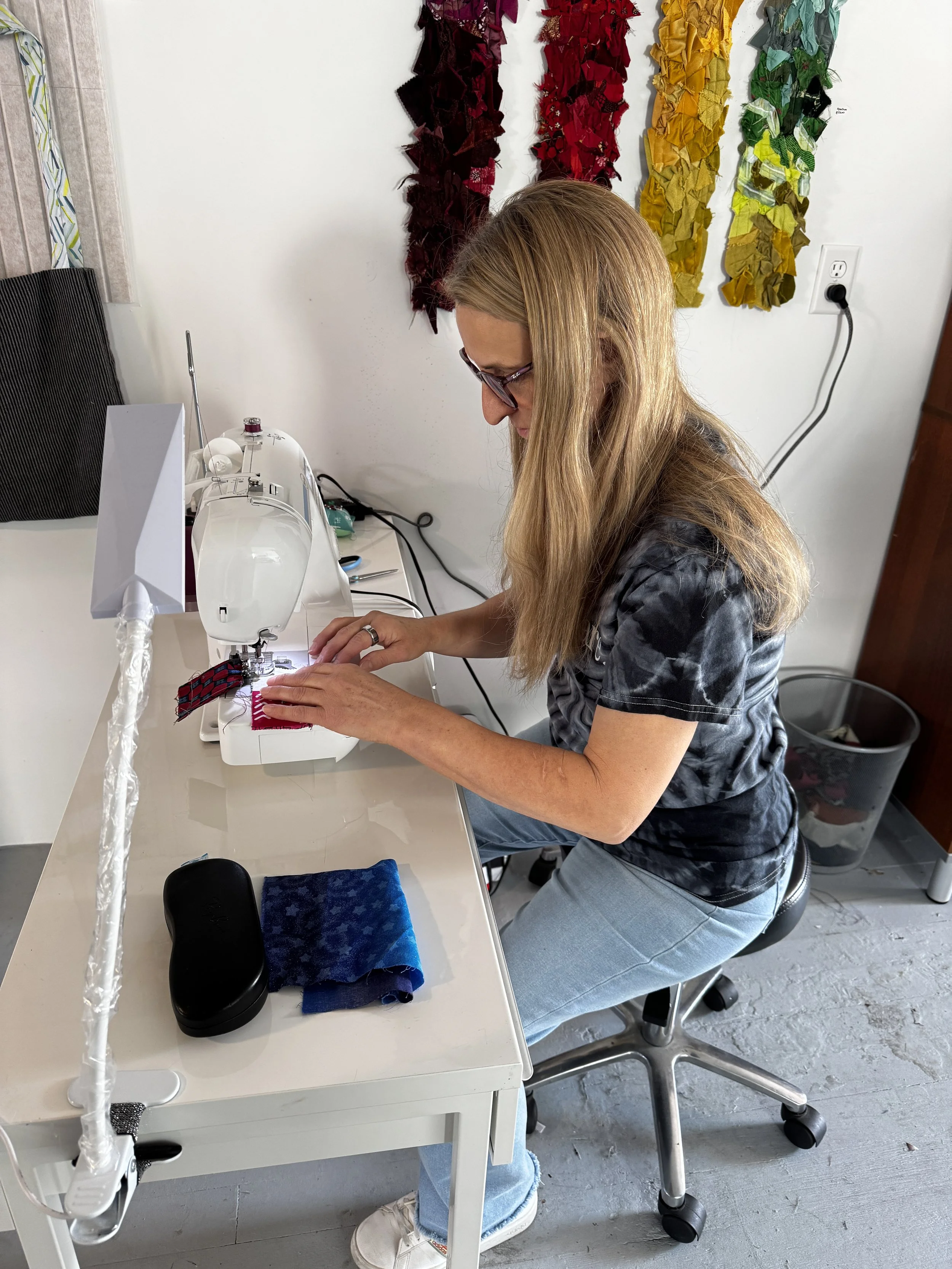 Woman sewing fabric on a sewing machine in a crafting room with colorful fabric decorations on the wall.