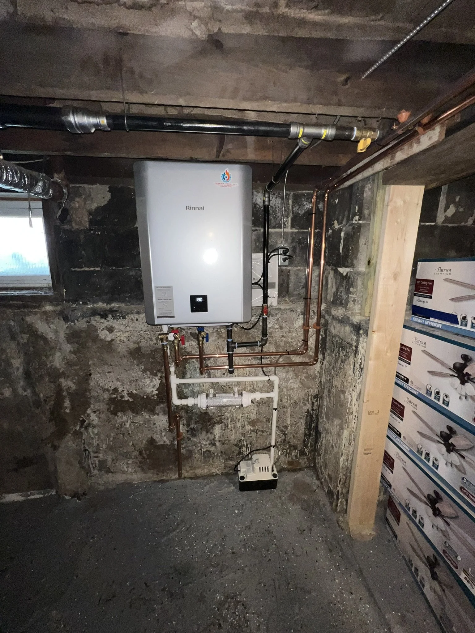A Rinnai tankless water heater mounted on a rough stone wall basement with copper and white PVC pipes connected, a small window on the left, and boxes of ceiling fans on the right.