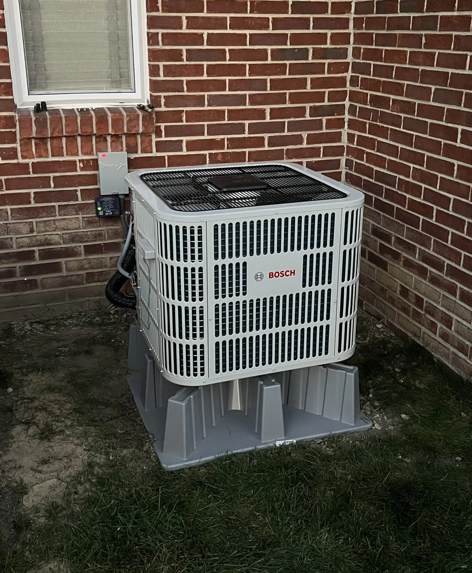An outdoor air conditioning unit with the Bosch logo, mounted on a concrete pad next to a brick house wall.